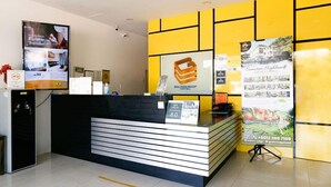 Ein Empfangsschalter mit modernem Design, bestehend aus Schwarz-Weiß-Farben und einem Logo. Hinter dem Schalter befinden sich gerahmte Zertifikate an der Wand, und ein großes Plakat wirbt für ein Hotel.