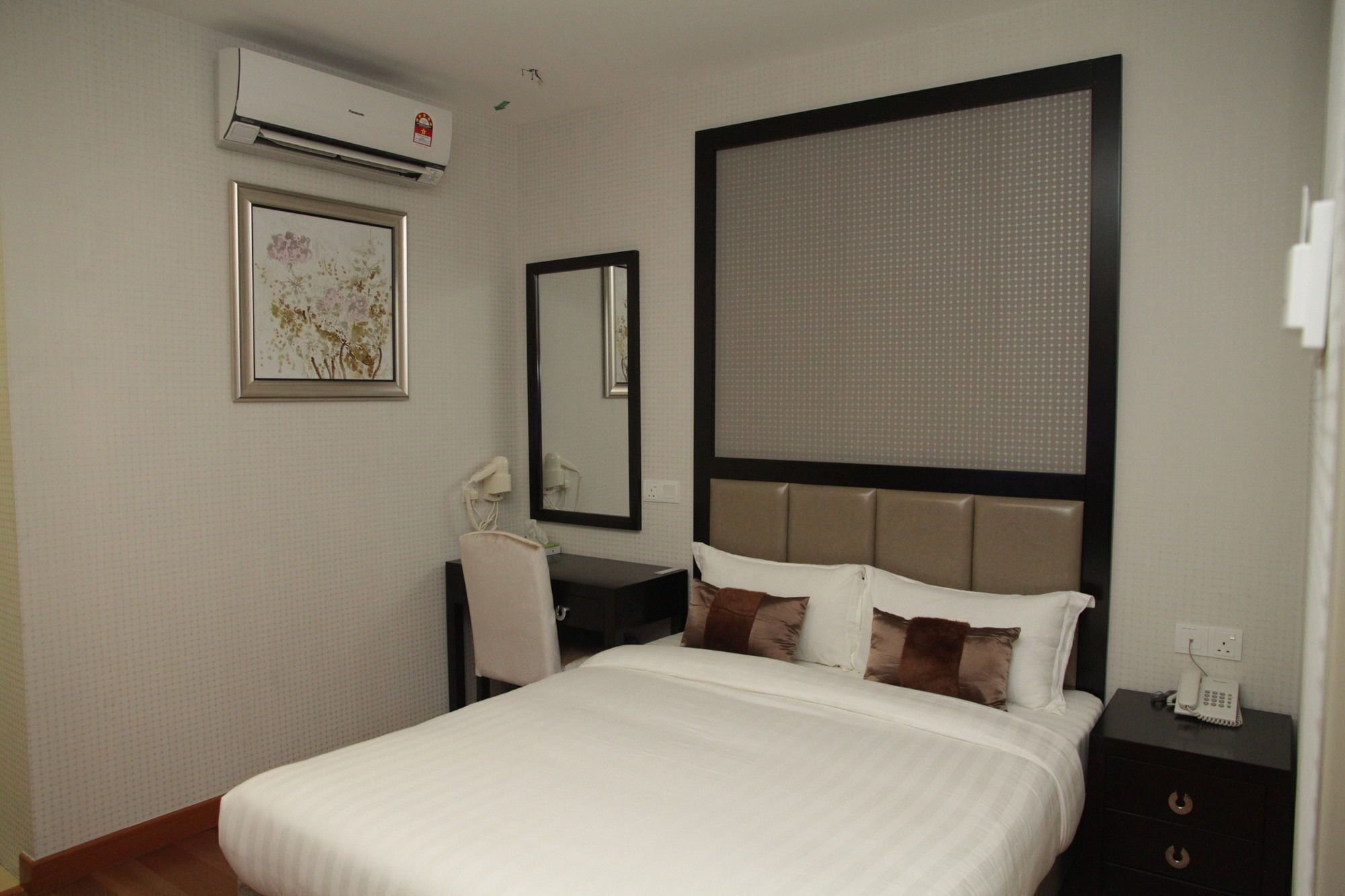 A hotel room with a bed, a desk with a chair, a mirror, and an air conditioning unit.