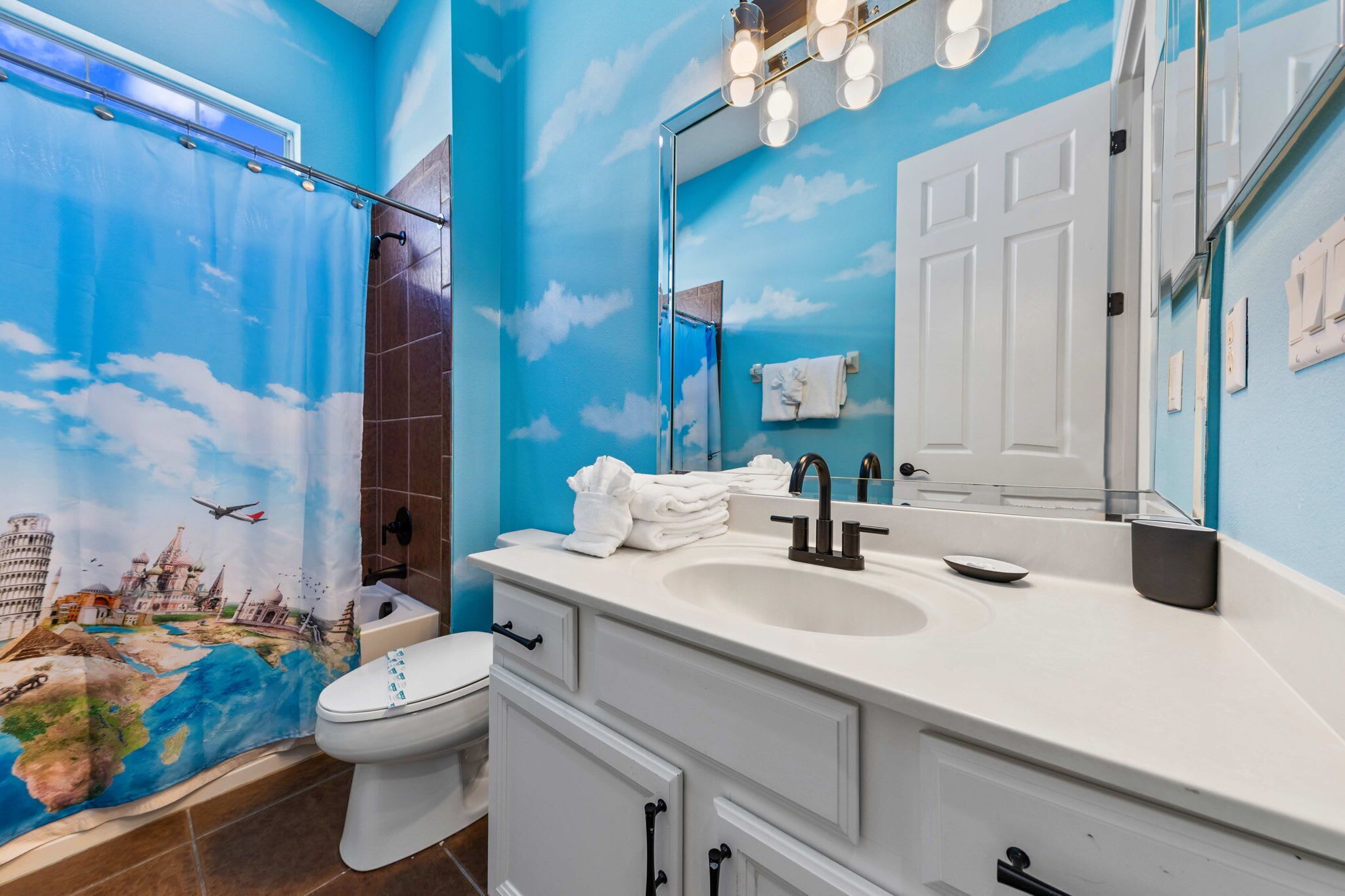 Une salle de bain avec une fresque représentant un ciel bleu et des nuages, un lavabo blanc et un miroir surmonté de lumières.