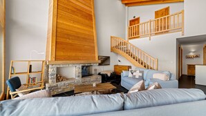 A cozy living room with a stone fireplace, a blue sofa, a wooden staircase, and a bookshelf. - Crevasse 07 - Elegant duplex & private sauna (Haute-Nendaz)