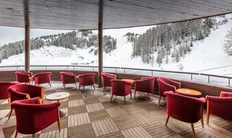 A covered outdoor seating area with red chairs and round tables, overlooking a snowy mountain landscape.