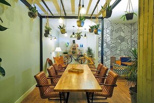 A dining area with a wooden table and wicker chairs, surrounded by hanging plants and a patterned wall.