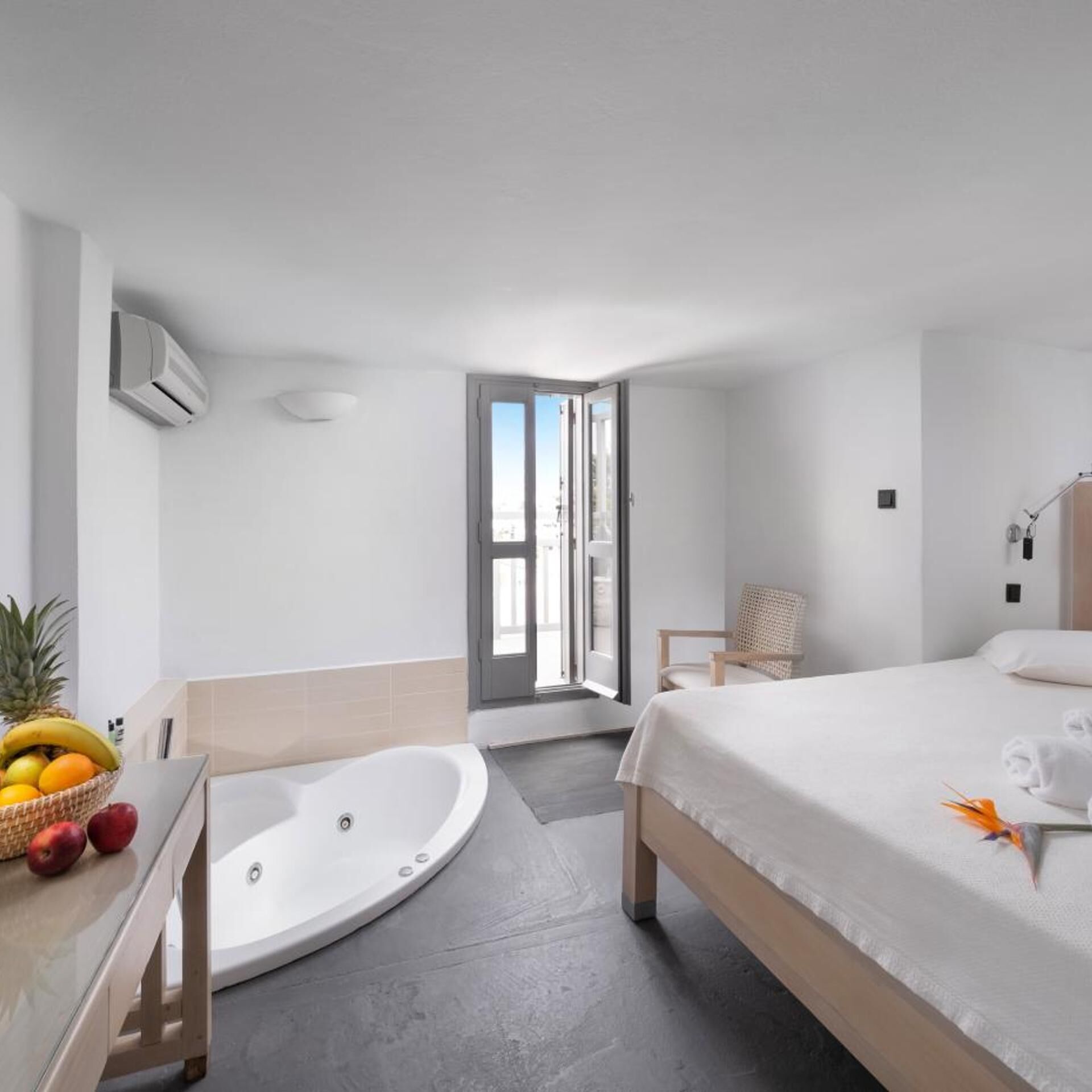 A modern hotel room with a large bed, a bathtub, a wicker chair, and a fruit basket.