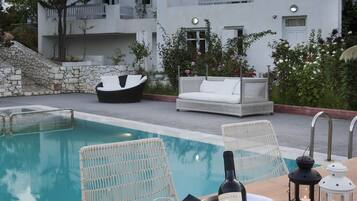 A poolside dining setup with a table set for two, a bottle of wine, and two glasses. There are lounge chairs and a white building in the background.