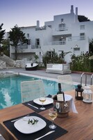 Montaje de comedor junto a la piscina con una mesa para dos, una botella de vino y dos copas. Se ven sillones de descanso y un edificio blanco al fondo.