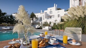 Une table de petit-déjeuner avec des assiettes, des tasses et un verre de jus d’orange, installée au bord d’une piscine, avec vue sur un bâtiment blanc.