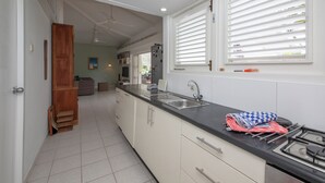 Eine Küche mit weißen Schränken, einer schwarzen Arbeitsplatte, einer Spüle und einem Herd. Es gibt ein Fenster mit Jalousien, einen Deckenventilator und eine Tür zu einem anderen Raum.