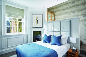 A bedroom with a large bed, a nightstand, a framed artwork, a window with a view, and a patterned wall. - Barnett Hill (Guildford)