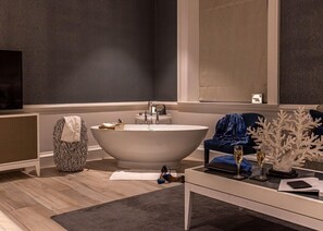 A modern bathroom with a freestanding bathtub, a vanity with a mirror, and a seating area with a blue chair and decorative items. - Barnett Hill (Guildford)