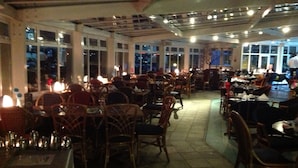 An indoor dining area with tables and chairs arranged in rows, large windows on one side, and a ceiling with exposed beams and lighting fixtures. - Little Haven Hotel (South Shields)