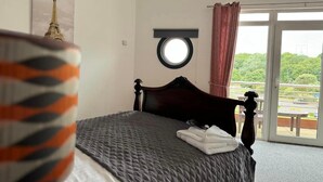A hotel room with a bed, a round mirror, a view of greenery and a road outside. - Little Haven Hotel (South Shields)