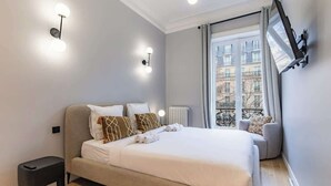 A modern bedroom with a large bed, a small stool, a radiator, and a window with a view of buildings. - Prestige Haussmannien & AC - Madeleine Paris 8 (Paris)