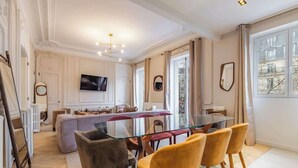 A spacious dining area with a glass table and modern chairs, a cozy seating area with a sofa and ottoman, and a large window offering a view of the outside. - Prestige Haussmannien & AC - Madeleine Paris 8 (Paris)
