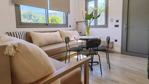 Ein modernes Wohnzimmer mit einem beigefarbenen Sofa, einem Esstisch mit Stühlen und großen Fenstern mit Jalousien.
