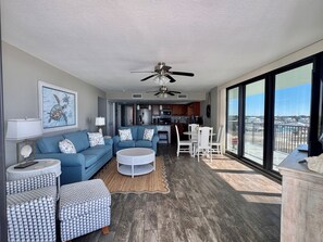 A spacious living area with a blue sofa set, a round coffee table, and a dining table with chairs. A ceiling fan is visible, and there is a large window offering a view of the outside. - Beachfront Condo, Pool, 700 sq ft Balcony!  Hot Tub, (Panama City Beach)