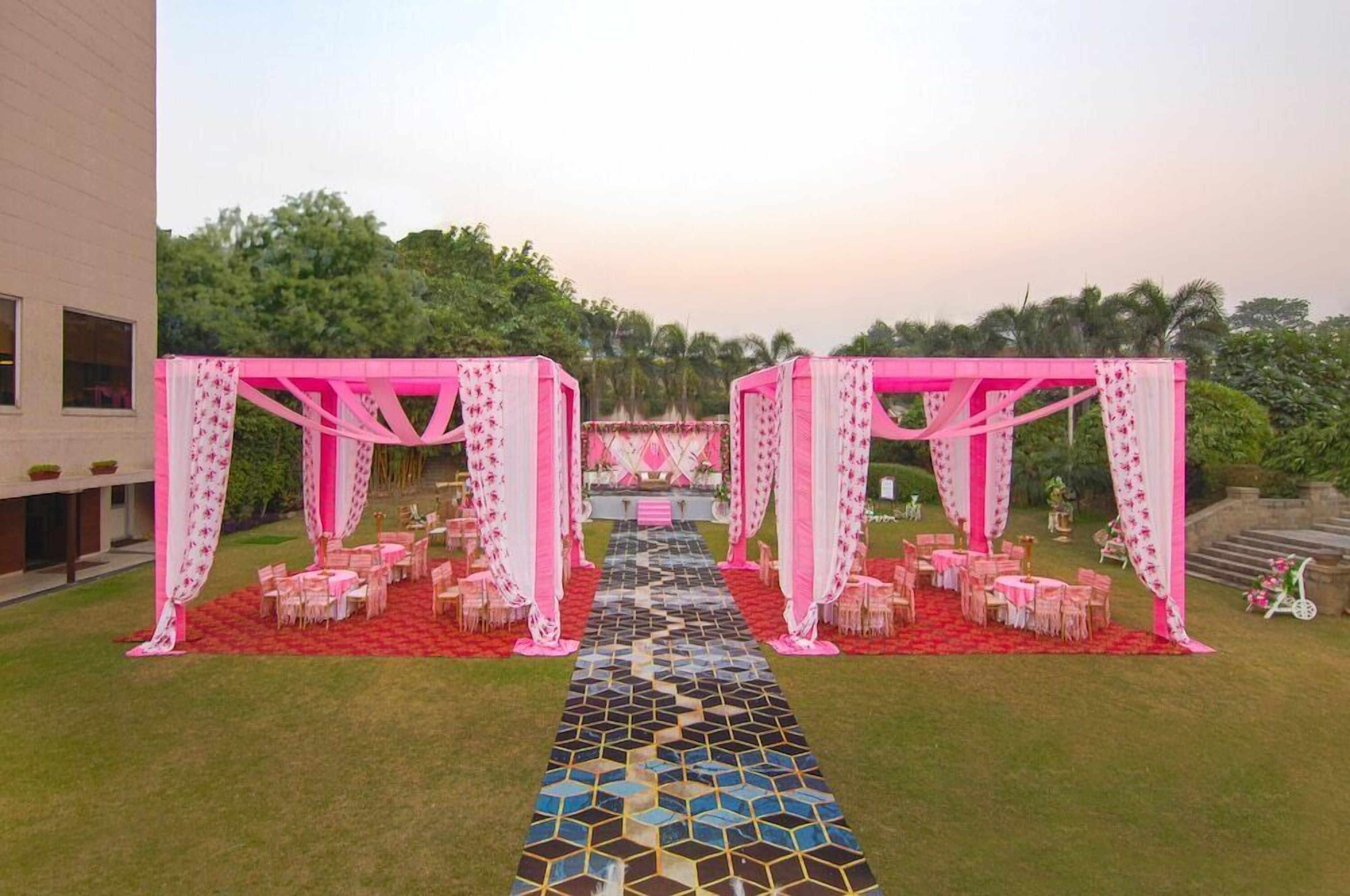A wedding setup with a decorated aisle, tables, and chairs, set against a building and greenery.