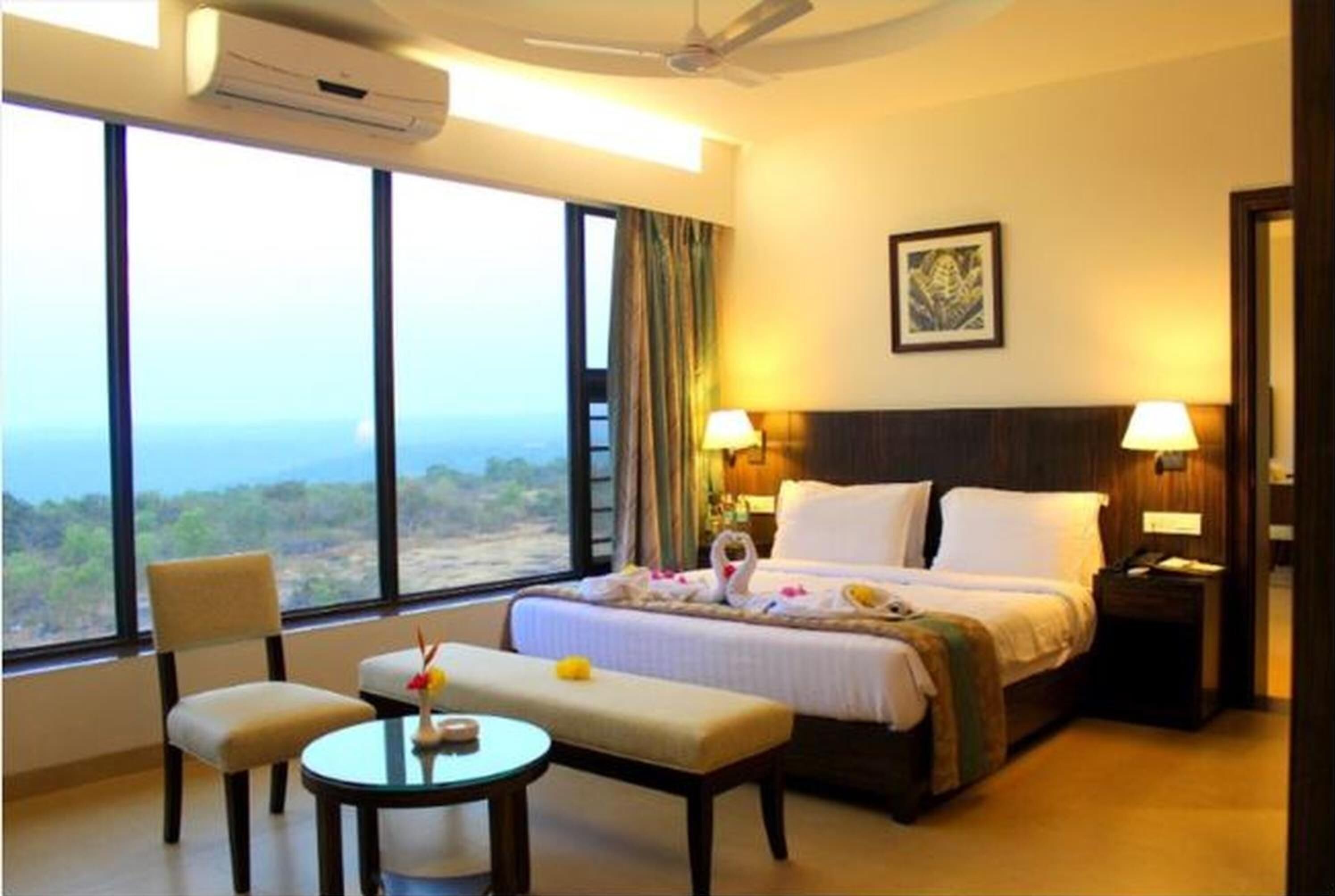 A hotel room with a large window, a bed, a bedside table, a chair, and a small table with a vase.