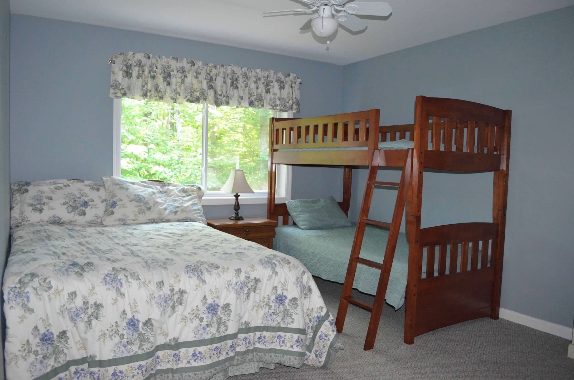 A bedroom with a bunk bed, a bedside table with a lamp, a window with a view of greenery, and a ceiling fan.