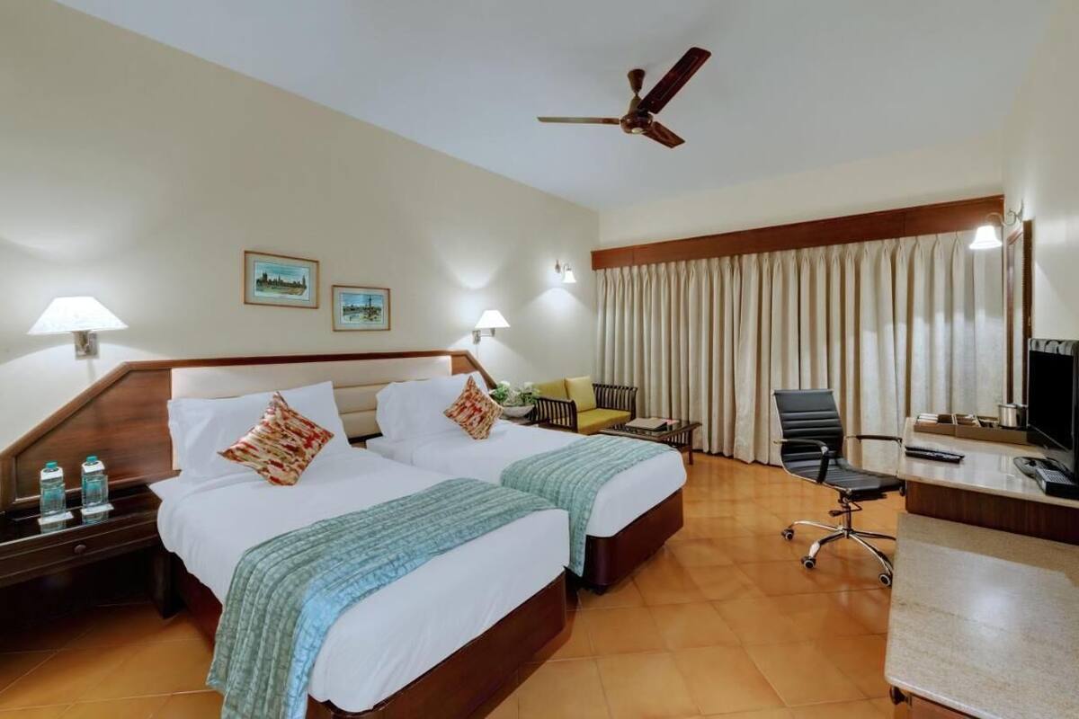 A hotel room with two beds, a desk with a computer, a chair, a TV, and a ceiling fan.