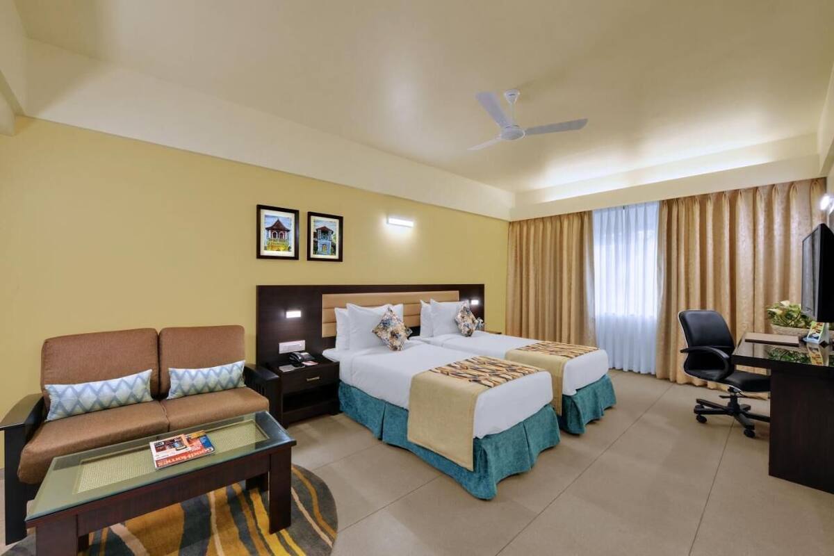 A hotel room with two beds, a sofa, a desk, a chair, a TV, and a ceiling fan.