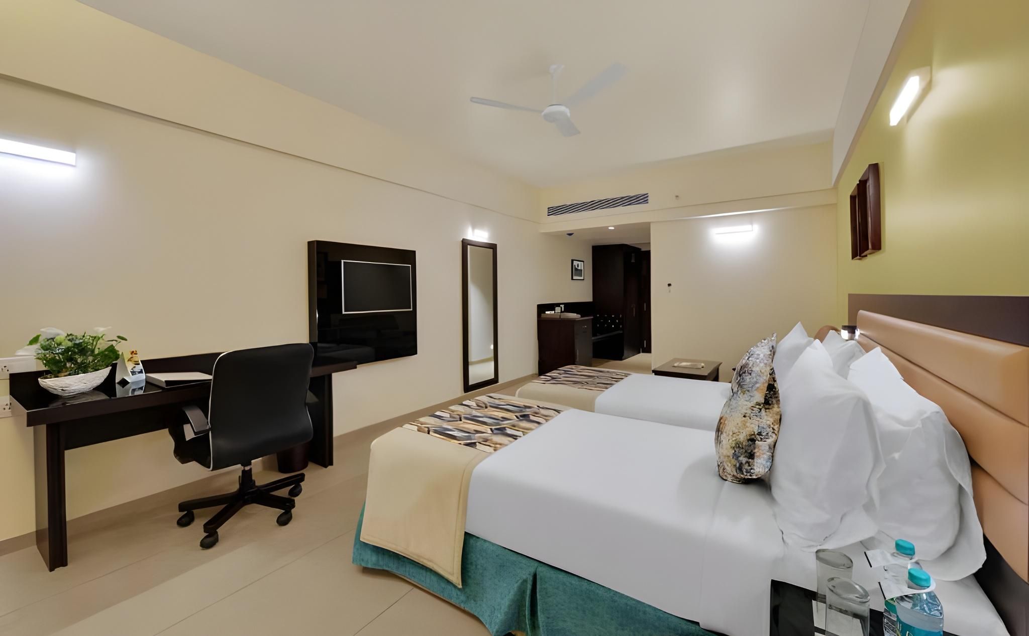A hotel room with a desk, two beds, a ceiling fan, and a TV.