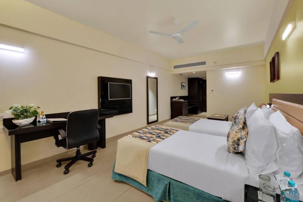 A hotel room with a desk, two beds, a TV, a ceiling fan, and a bathroom door.
