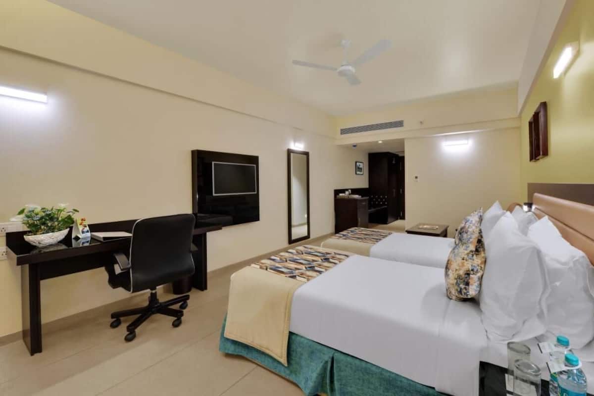 Habitación de hotel con escritorio, dos camas, televisión, ventilador de techo y puerta al baño.