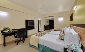 Habitación de hotel con escritorio, dos camas, televisión, ventilador de techo y puerta al baño.