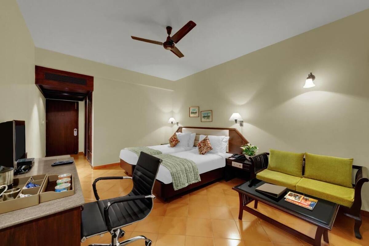 A hotel room with a bed, a desk with a computer, a chair, a sofa, a coffee table, a ceiling fan, and wall-mounted lights.