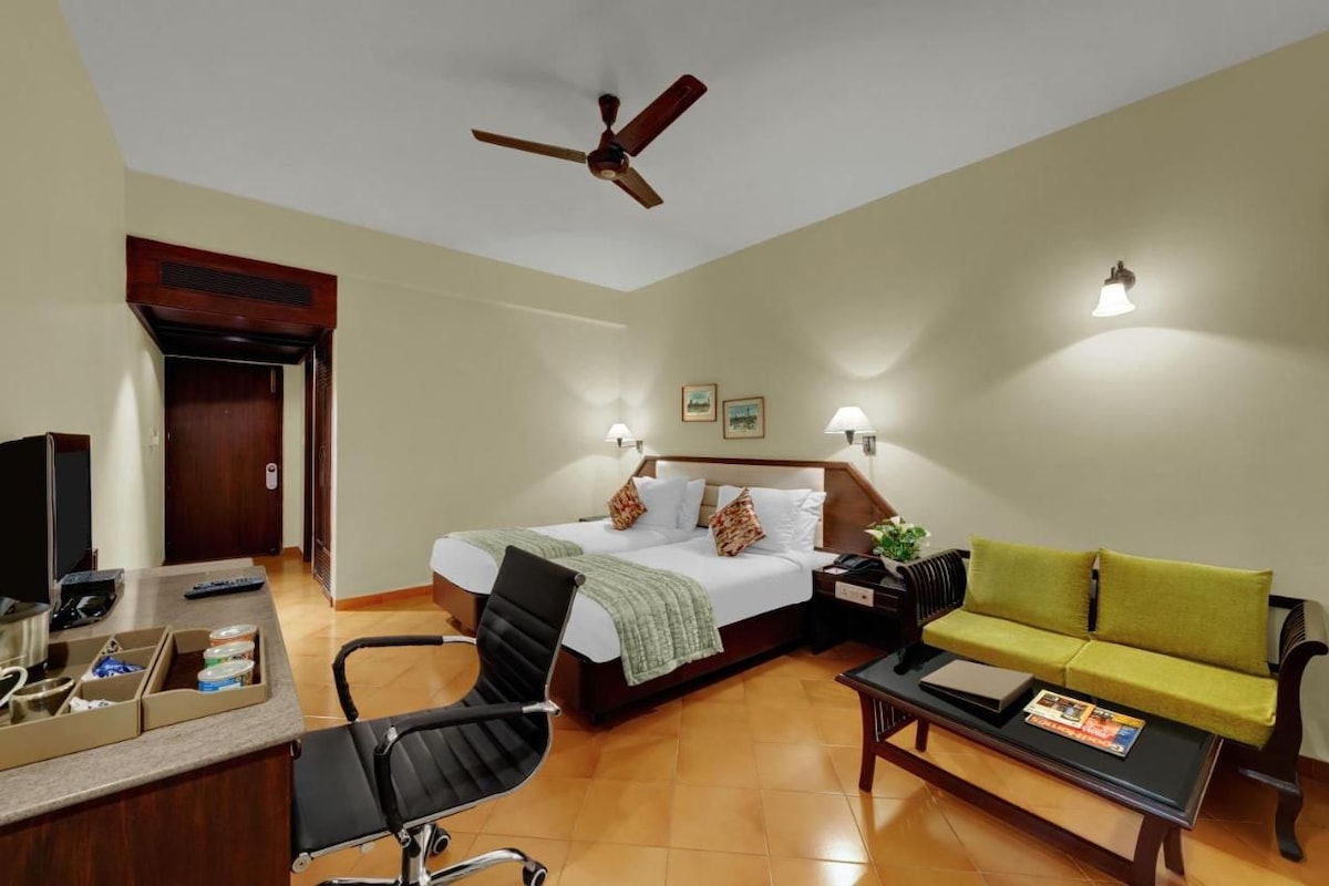 A hotel room with a bed, a desk with a computer, a chair, a sofa, a coffee table, a ceiling fan, and wall-mounted lights.