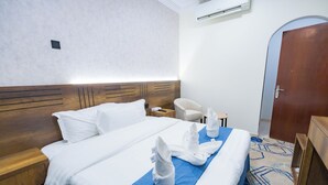 A hotel room with a wooden headboard, a large bed with white bedding, a blue accent on the bed, a wooden nightstand, a white chair, and a wooden wardrobe. - Asfour Hotel 03 (Makkah)