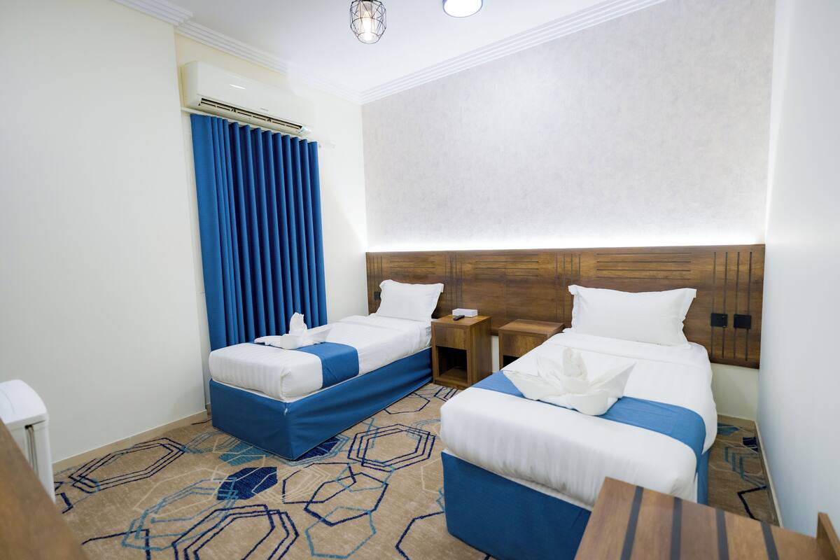 A hotel room with two beds, a wooden headboard, a blue curtain, and a patterned carpet.