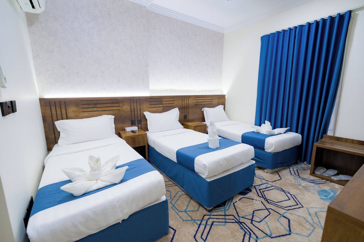 A hotel room with three beds, each with white linens and blue bedspreads. There are wooden headboards and a large window with blue curtains.