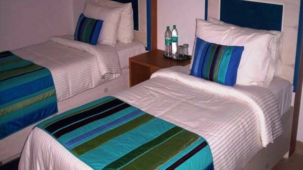 Une chambre d’hôtel avec deux lits, chacun doté de draps à rayures, une table de chevet en bois avec des bouteilles, et une tête de lit avec un panneau bleu.