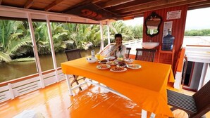 A dining area with a table set for a meal, surrounded by a wooden railing overlooking a body of water with palm trees. - Rimba Orangutan Eco Lodge (Kumai)