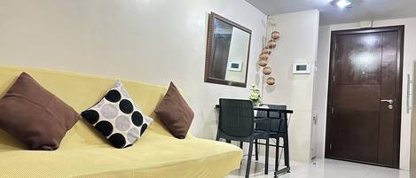 A living room with a yellow sofa, two brown pillows, a black chair, a small table with a vase, a mirror, and a door.