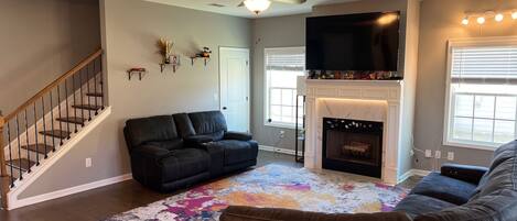 A living room with a fireplace, a black leather sofa, a colorful rug, and a staircase.