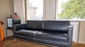 Een zwarte leren bank in een kamer met houten vloeren, een radiator en een raam met gordijnen.