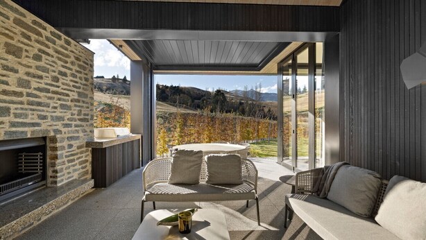 Un espace de vie extérieur moderne, agrémenté d’un mur en pierre, d’une cheminée intégrée, d’un coin salon avec une table et offrant une vue sur un paysage vallonné.