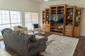 Ein Wohnzimmer mit grauem Sofa, zwei gemusterten Sesseln, einem Couchtisch und einer großen Holz-Unterhaltungsanlage mit Flachbildfernseher.