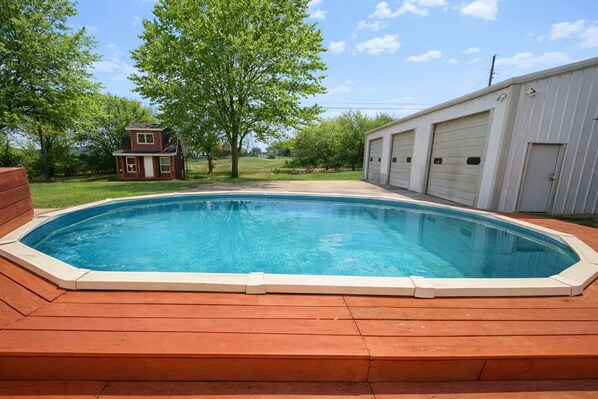 Ein rechteckiger Swimmingpool mit einer Holzdielen-Terrasse davor, umgeben von Bäumen und einem Gebäude mit Garage.