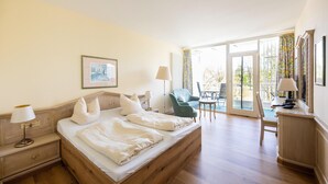 A hotel room with a wooden bed, two bedside tables with lamps, a chair, a desk, and a balcony with a view. - Liebevoll Eingerichtetes Studio am Kurpark in Bad Birnbach (Bad Birnbach)