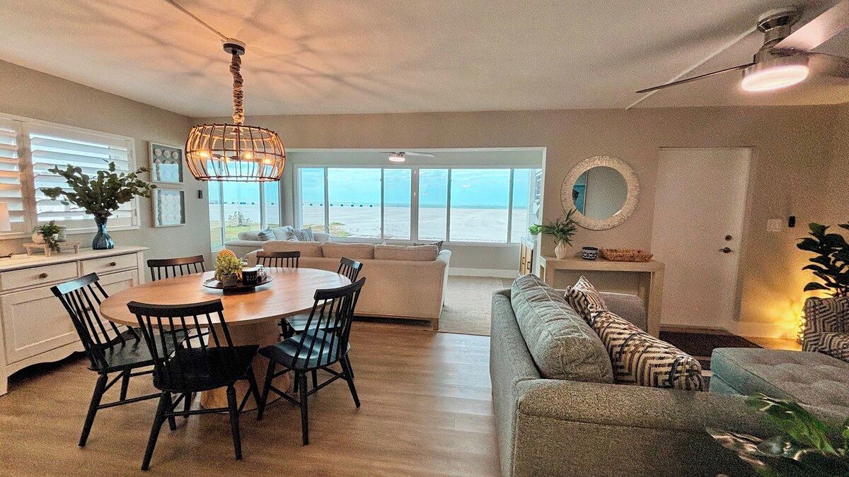 Een ruime woonkamer met een eettafel, stoelen, een bank en uitzicht op het strand.