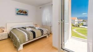 A bedroom with a bed, a nightstand, a lamp, a painting, and a view of a residential area through a glass door. - Newly decorated apartment in a quiet part of Baška (Baška)