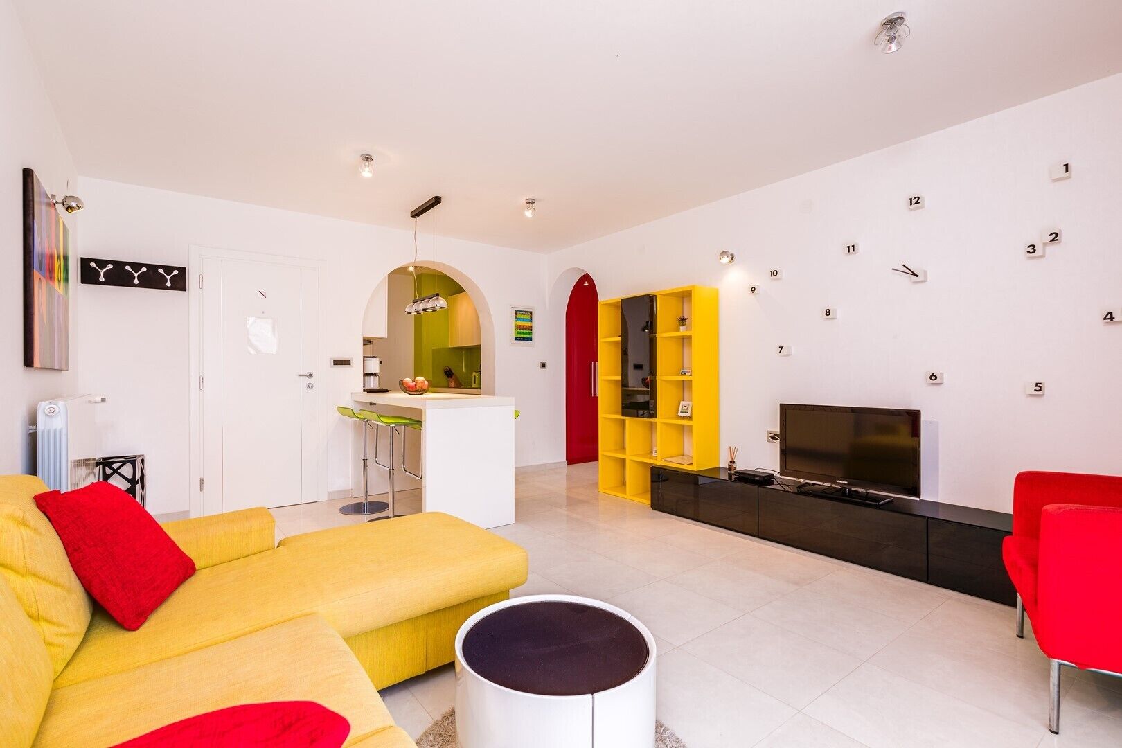 A modern living room with a yellow sofa, red cushions, a TV, and a yellow bookshelf.