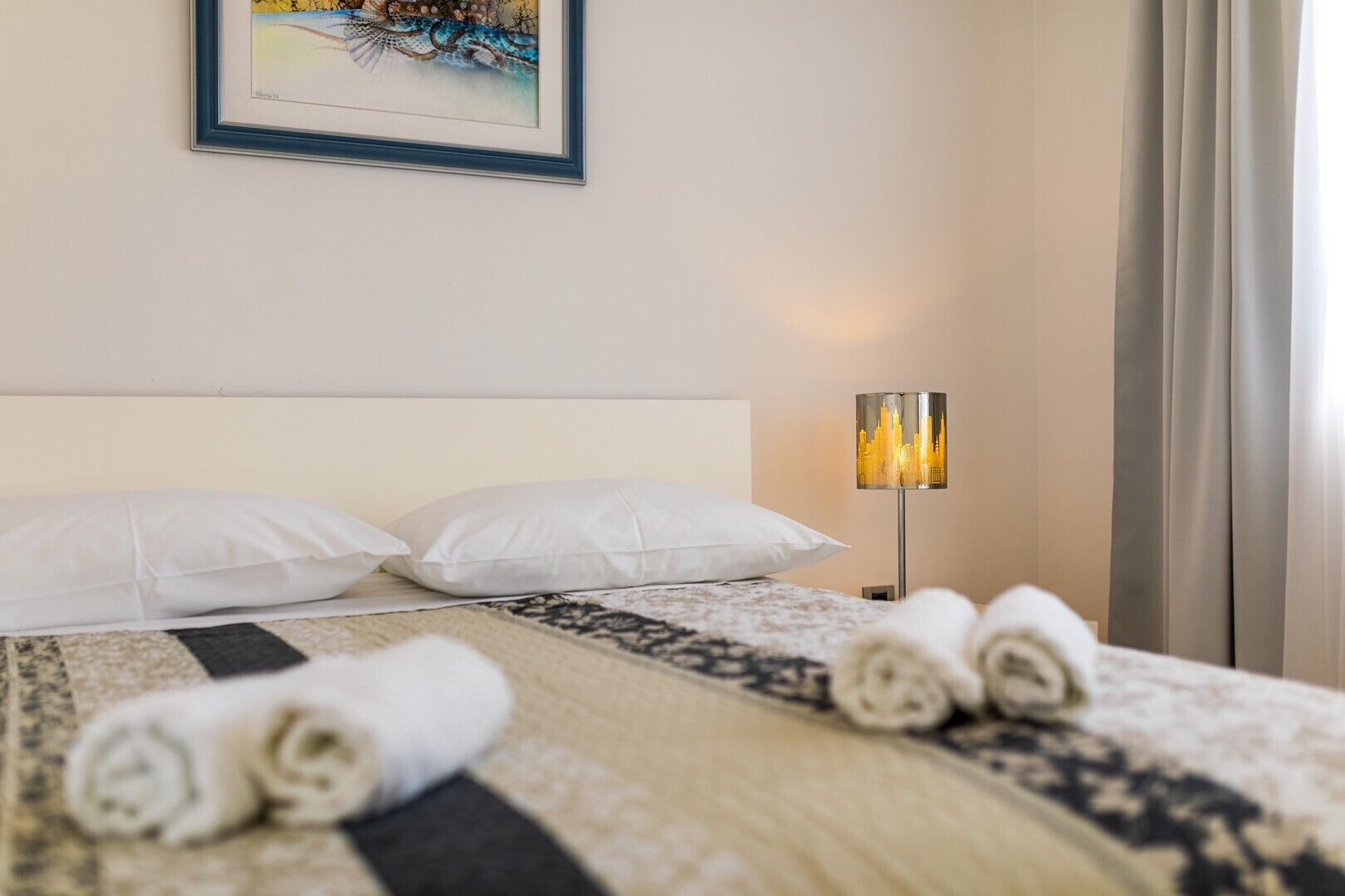 Ein ordentlich bezogenes Bett mit weißen Kissen und einer gemusterten Tagesdecke, ein gerahmtes Kunstwerk darüber und eine Stehlampe mit Stadtansicht-Motiv.