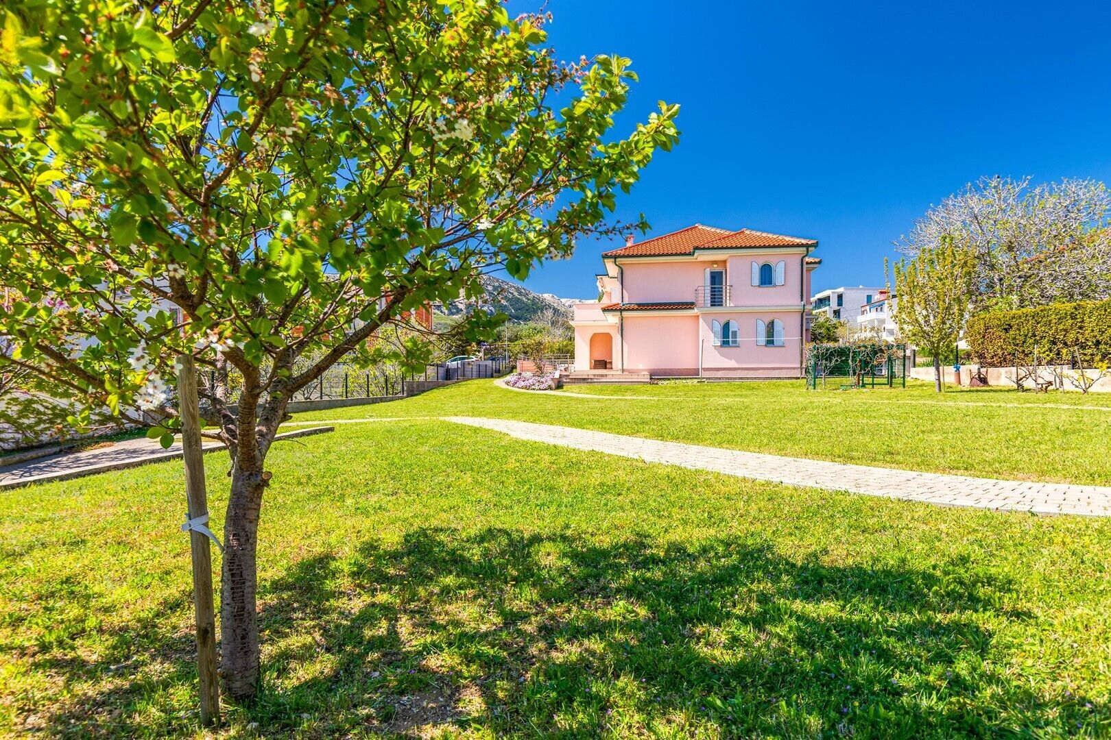 A pink villa with a tiled roof, surrounded by a well-maintained lawn and a paved pathway.