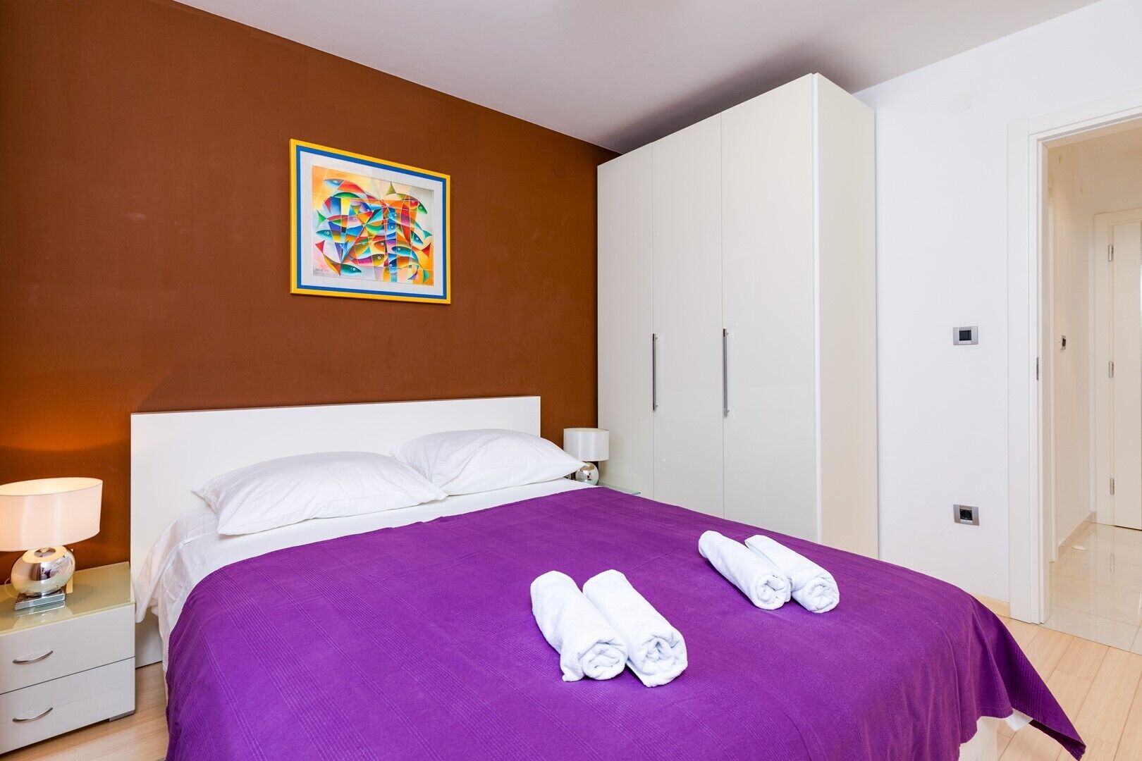 Habitación de hotel con cama, mesita de noche, lámpara, armario y un cuadro en la pared.
