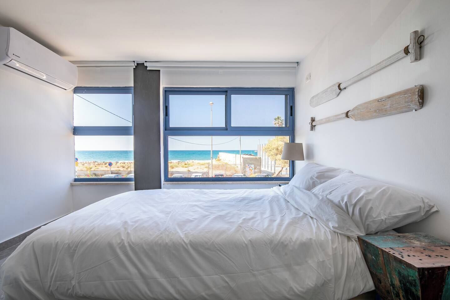 Un dormitorio con cama, ventana con vista a la playa y una lámpara fijada a la pared.