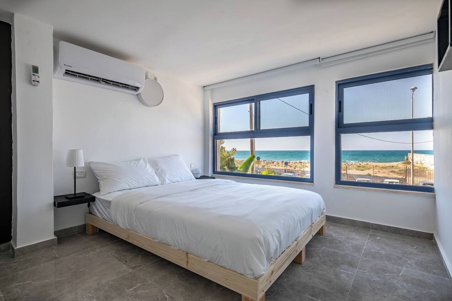 Une chambre avec un lit, une table de chevet, un appareil de climatisation et deux fenêtres donnant sur la plage.